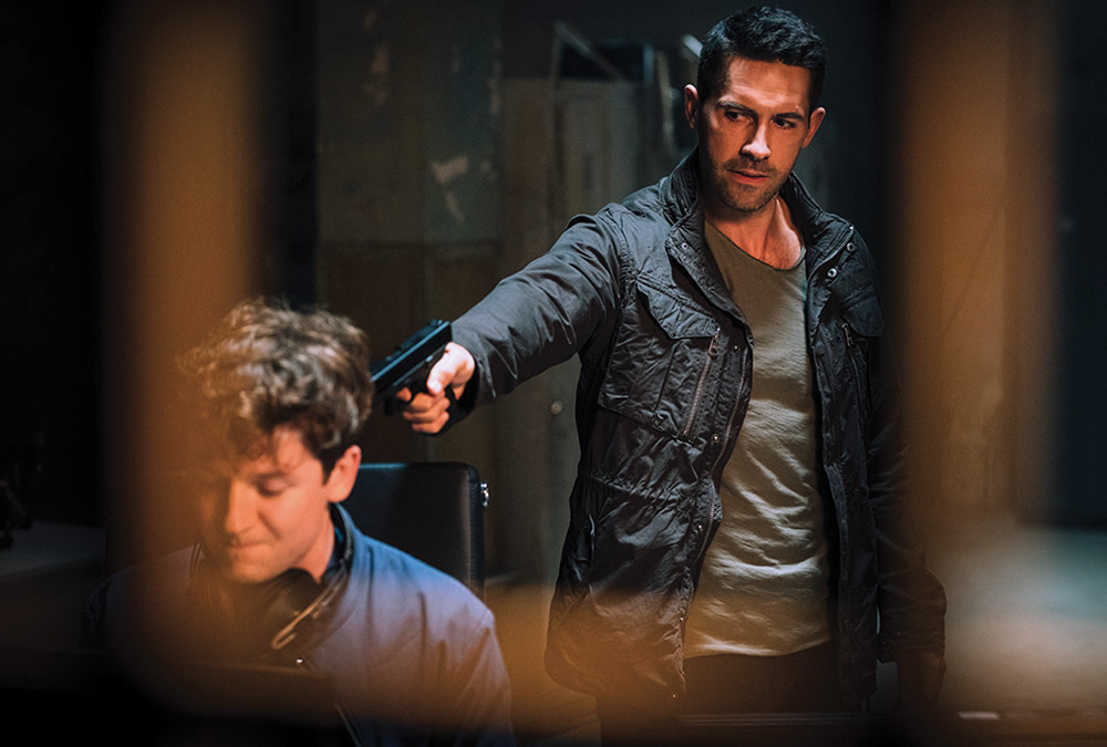 A imagem mostra o protagonista Martin Baxter apontando uma arma na cabeça de uma pessoa rendida, ajoelhada e de costas para ele. Cena do filme Legacy of Lies.