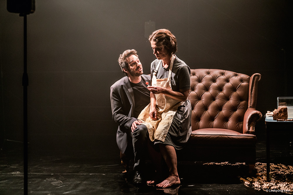 Uma mulher de avental sentada em um sofá ouve um homem agachado ao seu lado, em um palco de teatro