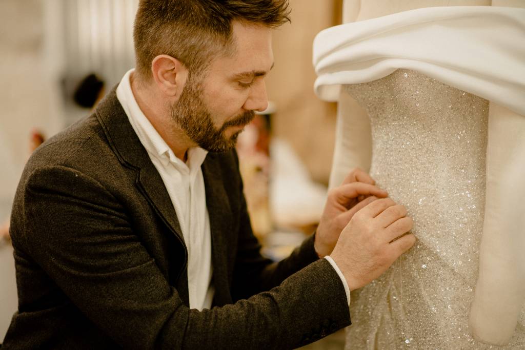 Lucas Anderi costura trecho de vestido de noiva na imagem. Aparece de paletó preto e camisa branca.