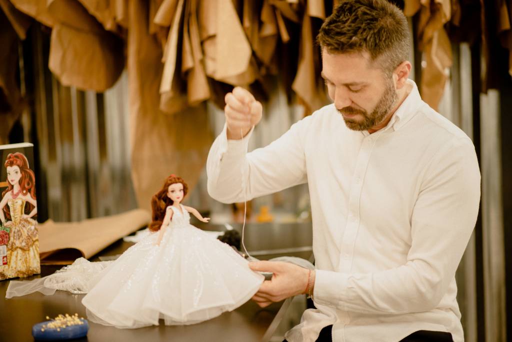 Lucas Anderi aparece costurando vestido de noiva em boneca. Veste camisa branca e calça preta.