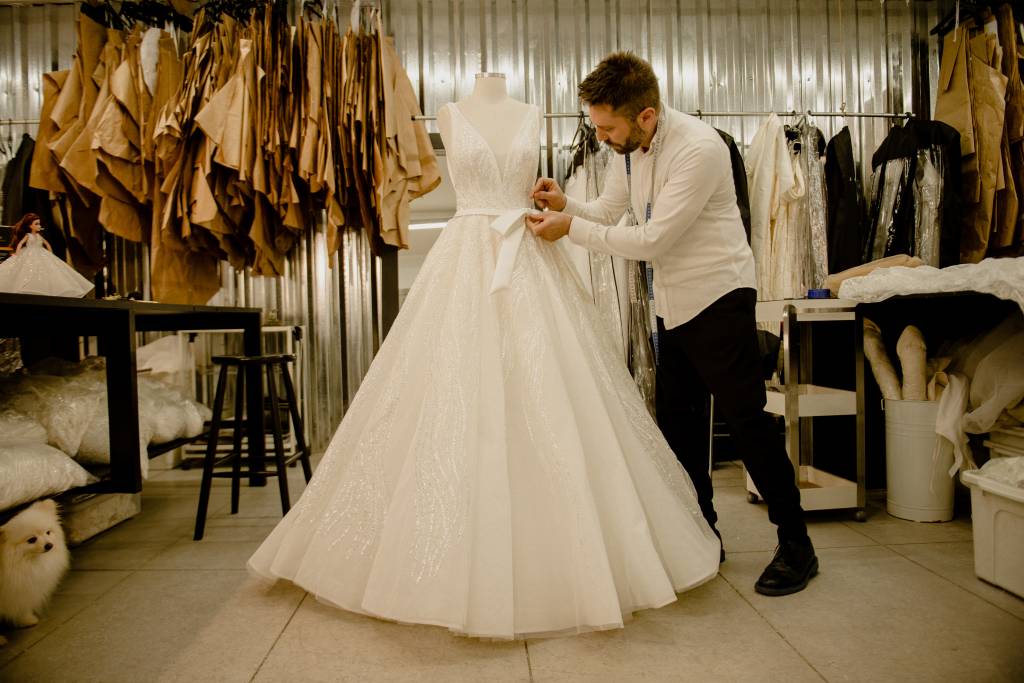 Lucas Anderi costura trecho de vestido de noiva na imagem. Aparece de paletó preto e camisa branca ao lado do vestido em seu ateliê.