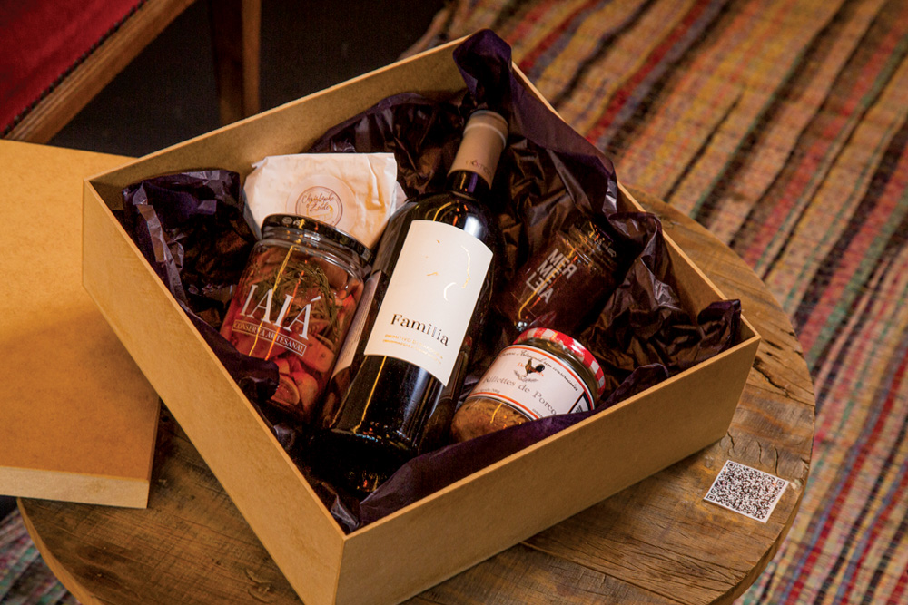 Em uma caixa de madeira decorada com papel de seda marrom, estão uma garrafa de vinho, três potes de vidro e um queijo embalado em papel de cor branca.