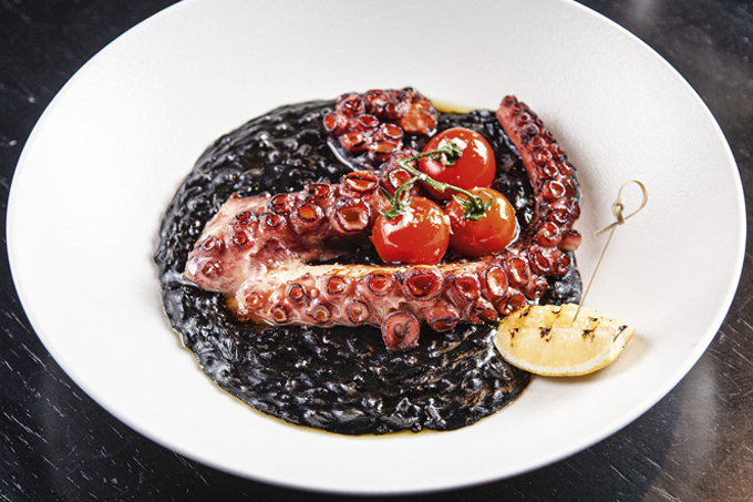 Prato fundo com arroz de tinta de lula coberto por tentáculos de polvo e tomatinhos em rama.