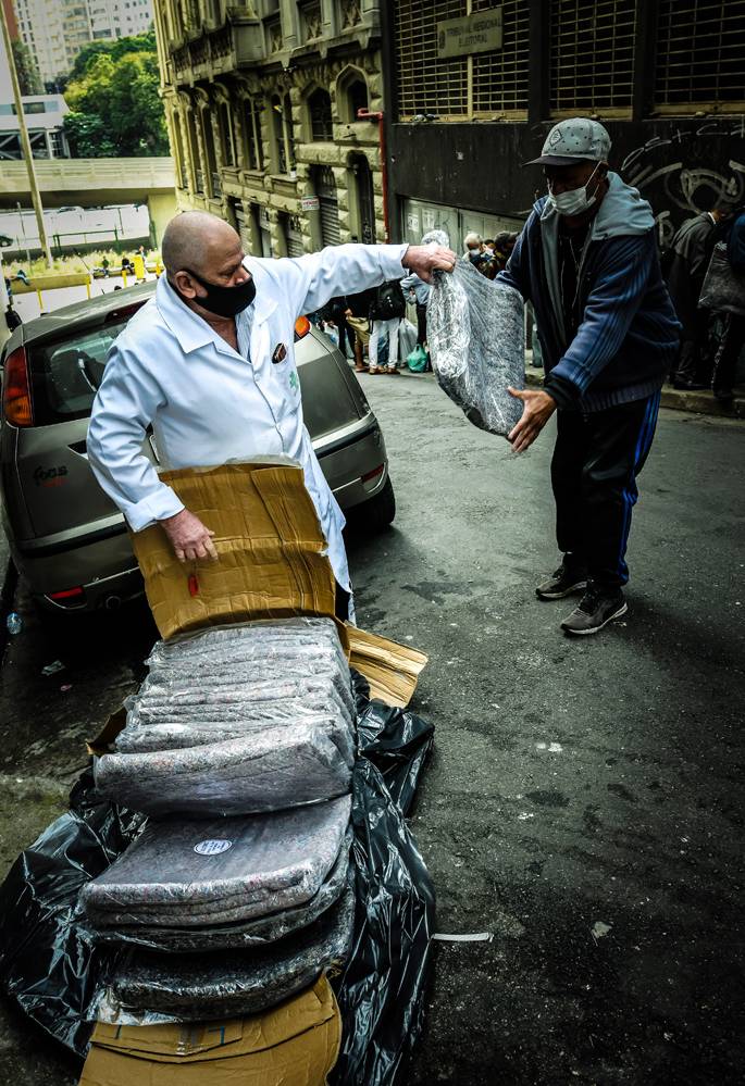 A imagem mostra Robson, com um carrinho cheio de cobertores, entregando um deles à outra pessoa na rua
