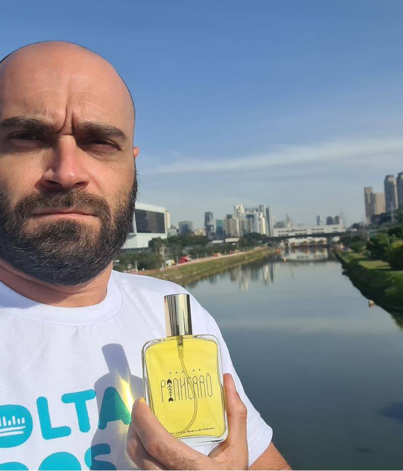 Marcelo Reis, porta-voz do movimento Volta Pinheiros, segura frasco de líquido amarelo em frente ao Rio Pinheiros. Ele é careca e tem barba.