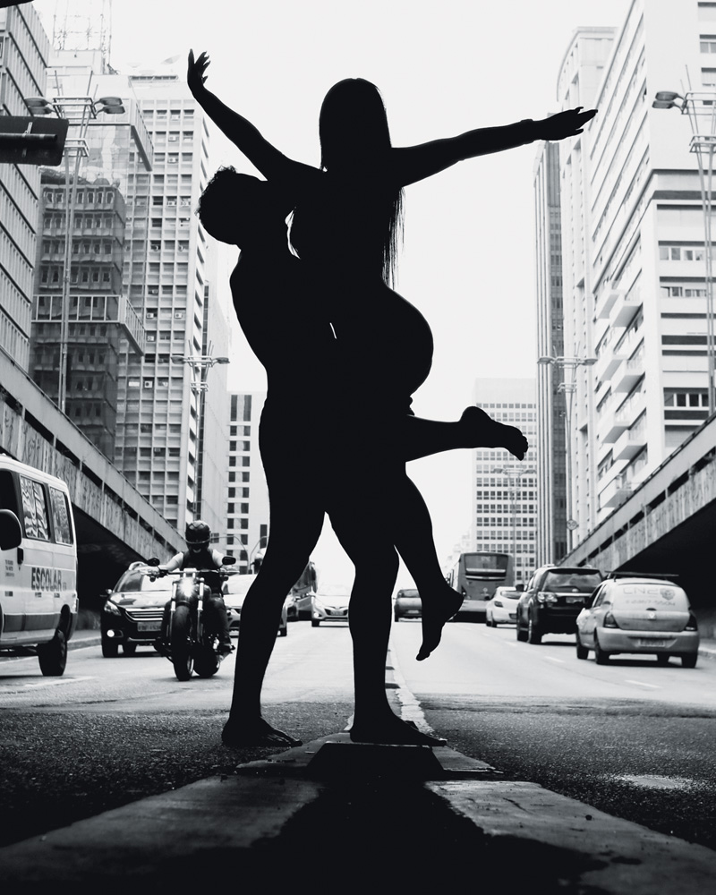 foto em preto e branco da silhueta de arthur e luana posando nus para a foto na paulista. eles estão no meio da rua, com a avenida ao fundo, fazendo uma pose em que ele a segura, enquanto ela abre os braços
