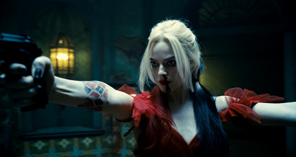 A imagem mostra Margot, vestida de Arlequina em cena do filme, empunhando uma arma em cada mãos apontando para diferentes direções. Seu nariz está sangrando e ela está com uma expressão de seriedade