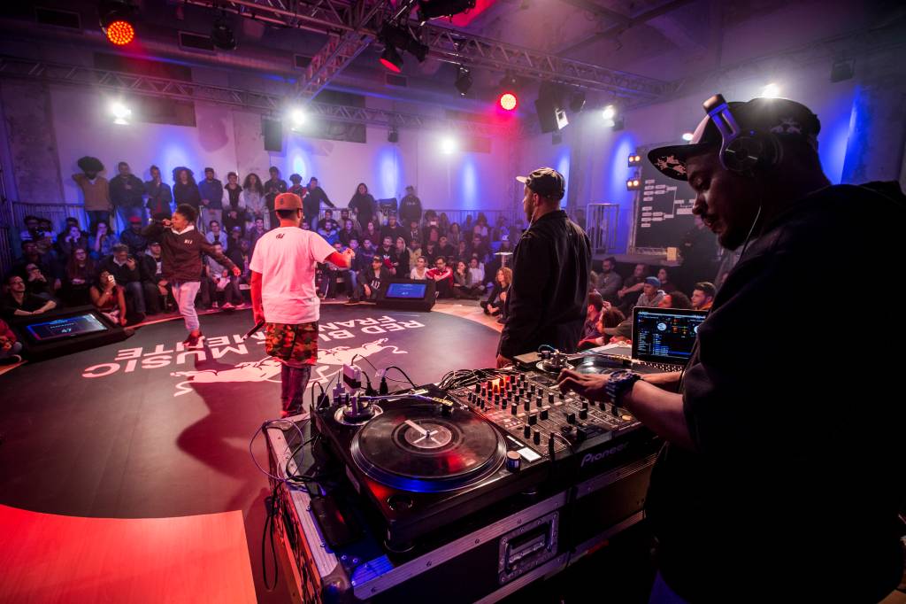 Foto mostra palco escuro com holofotes na parte de cima. No meio, dois rappers batalham com rimas e poesias faladas. Ao fundo, uma plateia assiste o evento. Em primeiro plano, um DJ aparece controlando sua mesa de mixagem.