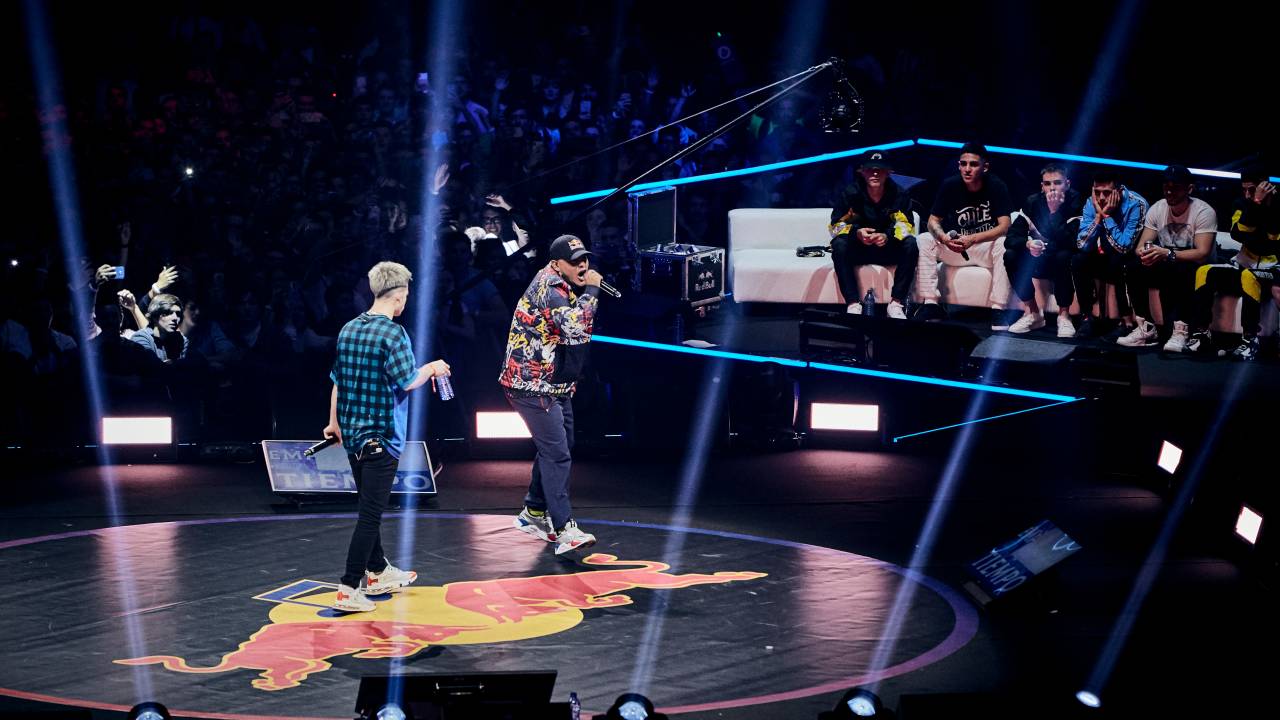 Foto mostra palco escuro com holofotes voltados para cima. No meio, dois rappers batalham com rimas e poesias faladas.