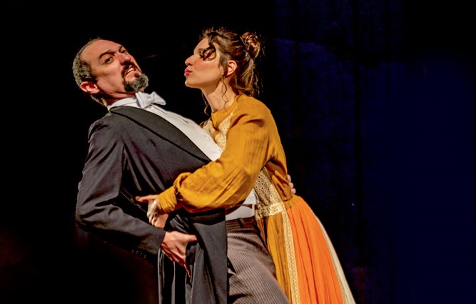 Em cena, em um palco de teatro, uma mulher com braços em volta de um homem, faz movimento de tentar beijá-lo. Ele se esquiva