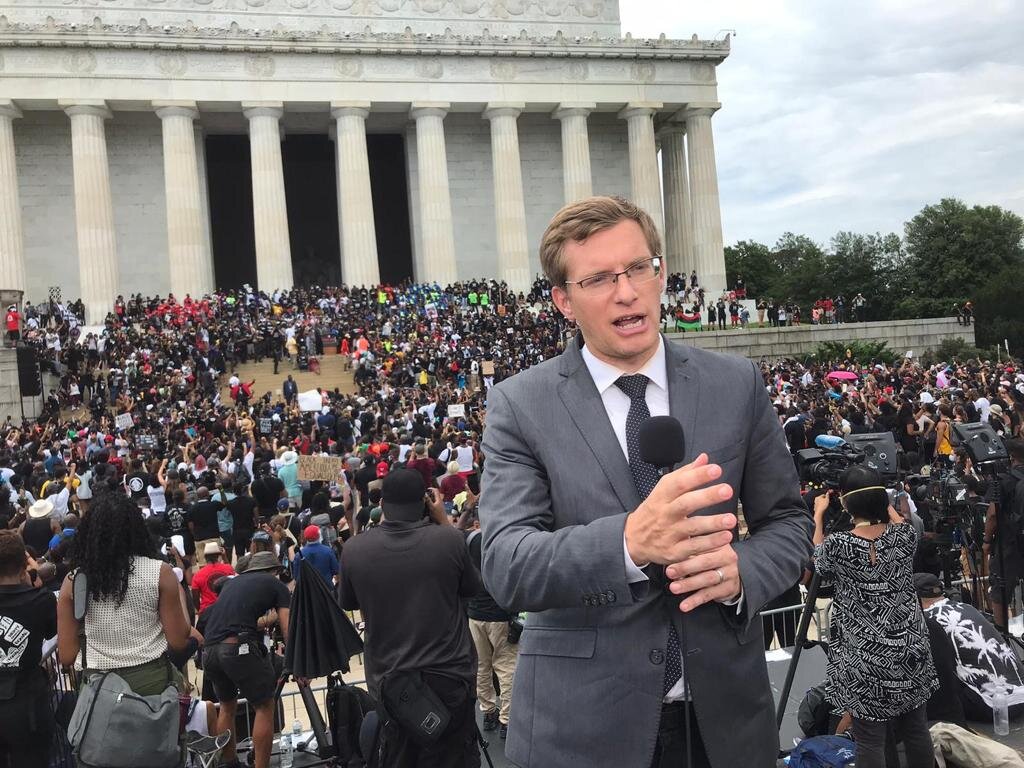 Philip Crowther aparece de terno falando em microfone. Ao fundo, multidão ocupa entrada do Capitólio dos Estados Unidos.
