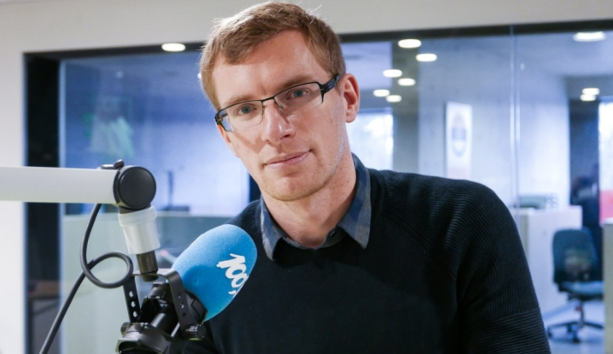 Philip Crowther aparece de suéter preto e óculos de grau em frente a microfone azul de rádio.
