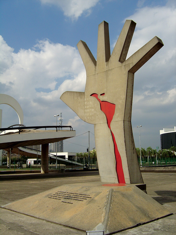 A imagem mostra a escultura na Praça do Sol, uma mão aberta elevada com um desenho vermelho na palma dela.