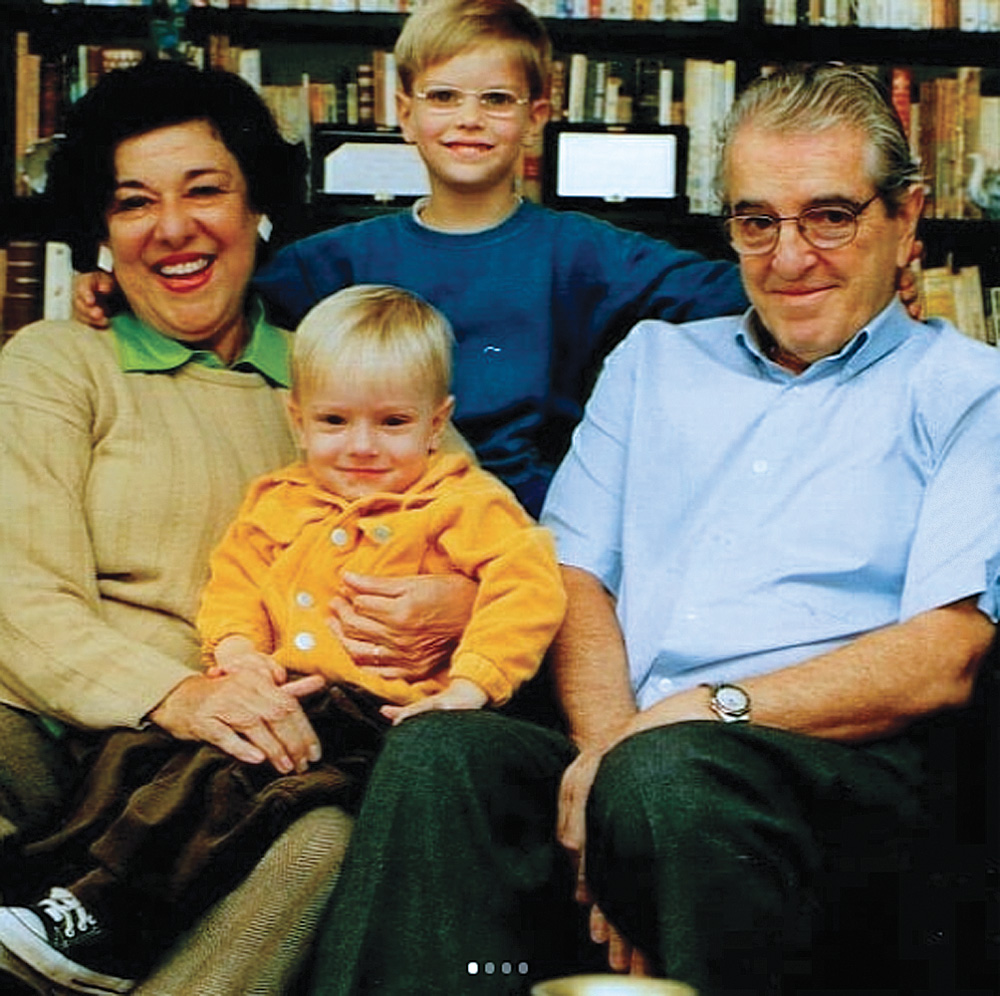 ruth rocha com seu marido, eduardo rocha, e seus netos pedro e miguel, sorrindo posando para a foto. pedro está sentado no colo de ruth, que está sentada ao lado de eduardo. miguel está de pé atrás deles com um braço nas costas de cada avô