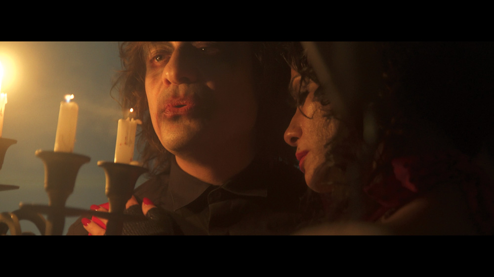Em uma cena escura, uma mulher se apoia no ombro de um homem com maquiagem pesada que está segurando um candelabro com velas acesas