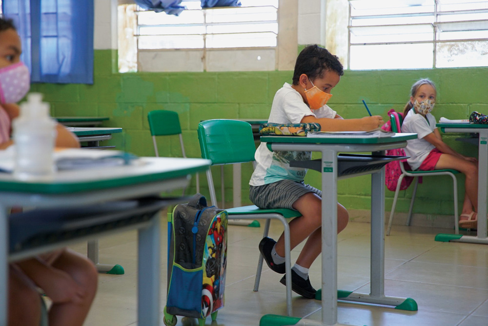 A imagem mostra alunos em uma sala de aula, distanciados, usando máscara enquanto escrevem em cadernos nas suas mesas.