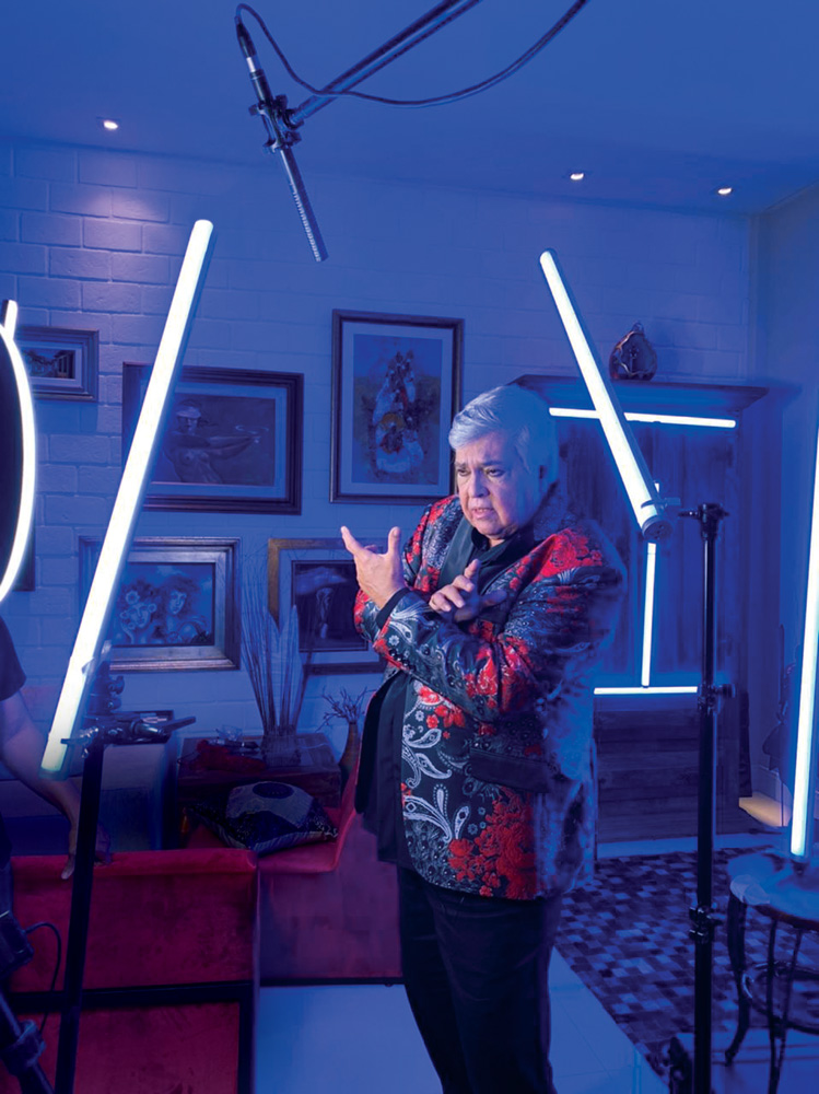 Sidney Magal (DIVULGAÇÃO) (1).jpeg Sidney Magal está de pé em sala iluminada com dois bastões azuis. Ele veste blazer azul e vermelho e ergue a mão esquerda em frente a uma câmera.