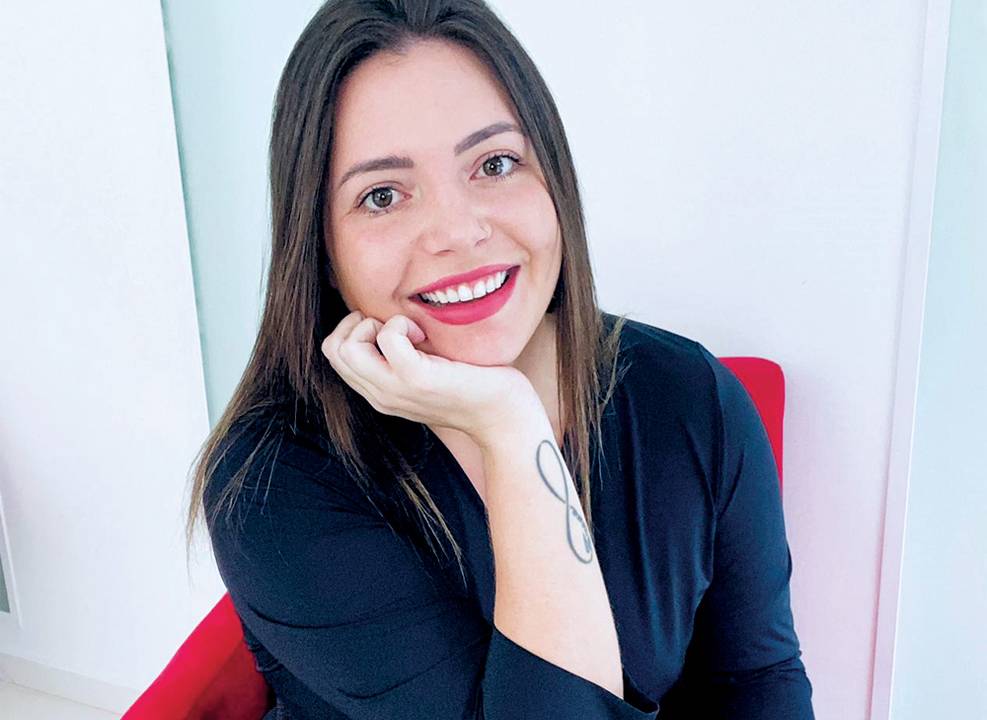 Stephanie Seitz sorri sentada com a mão no queixo. Tem cabelos escuros longos e veste roupas pretas. Está sentada em poltrona vermelha e em um local com fundo branco.