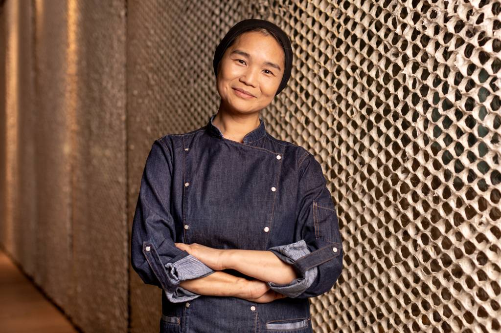 A chef Telma Shiraishi sorrindo de braços cruzados em frente à parede de textura rendada.