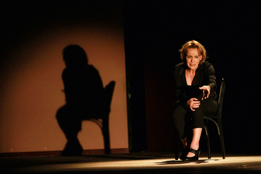 Mulher com expressão investigativa sentada em uma cadeira em um palco de teatro com luz baixa
