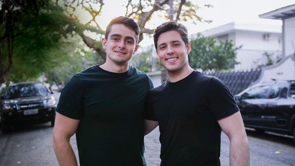 Cayo Syllos e Bruno Adami posam lado a lado sorrindo em rua arborizada. Os dois usam camisetas pretas, são brancos e têm cabelo castanho.