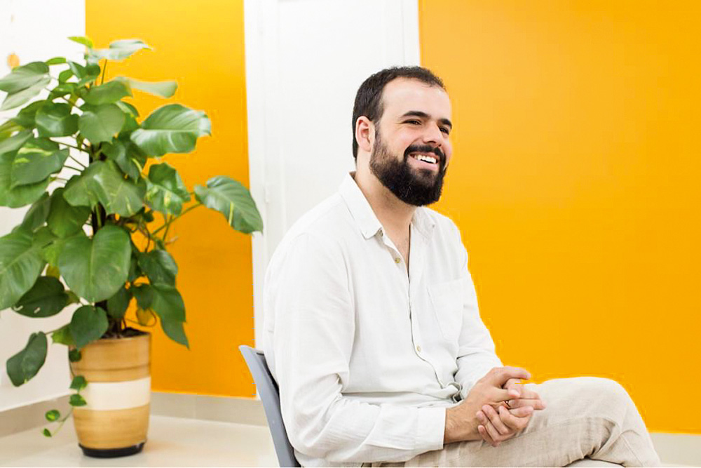 nickson gabriel sentado sorrindo com mãos juntas olhando para o horizonte. o fundo tem uma parede amarela e uma planta