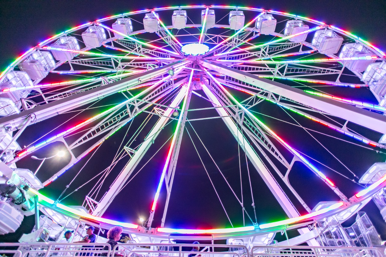 À noite, foto de baixo de uma roda-gigante girando. Tem luzes de LED coloridas em toda a estrutura do brinquedo
