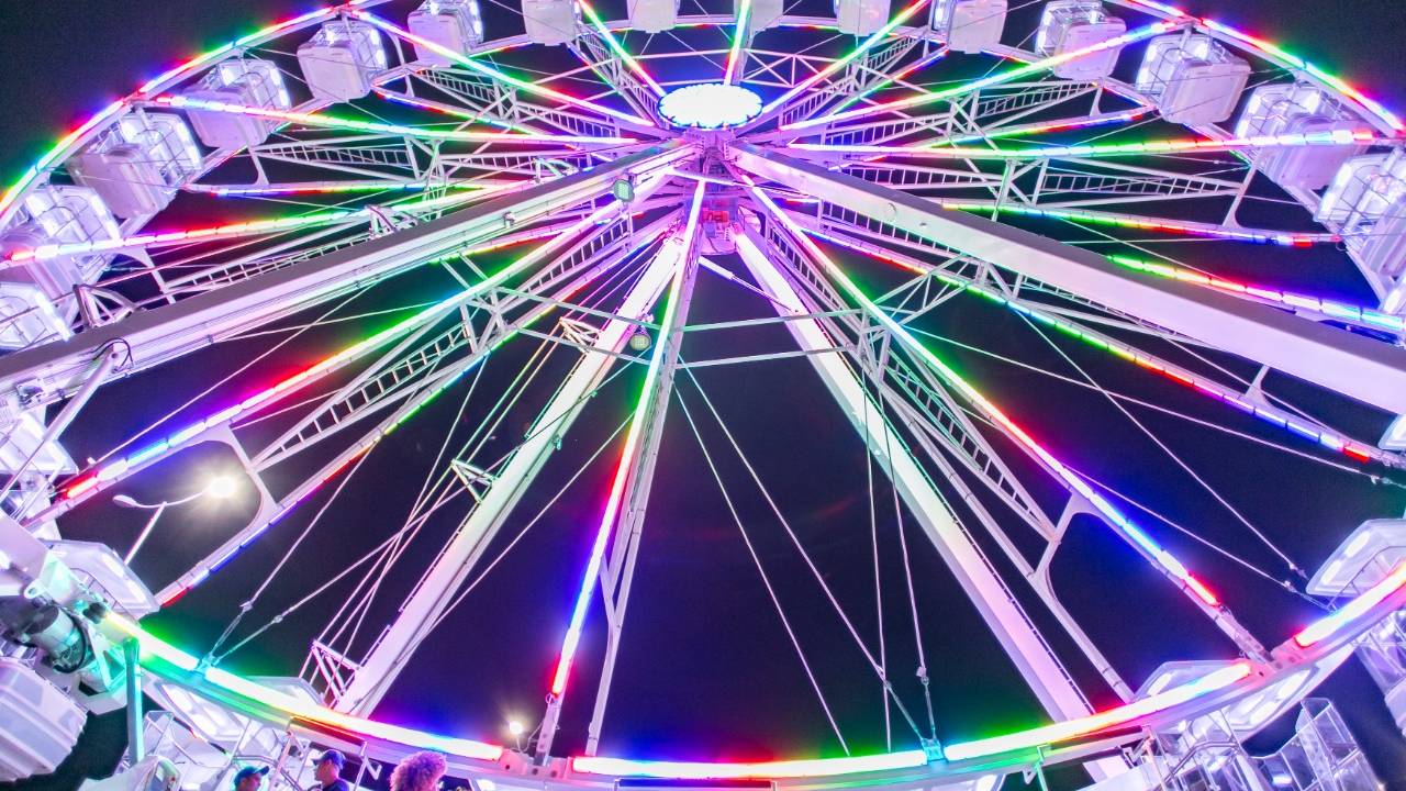 À noite, foto de baixo de uma roda-gigante girando. Tem luzes de LED coloridas em toda a estrutura do brinquedo