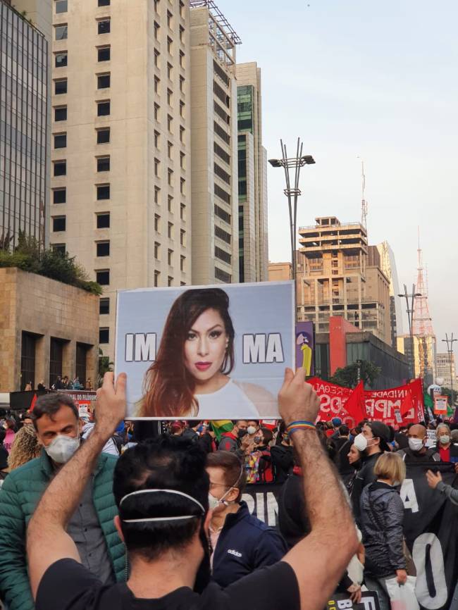 Manifestante segurando cartaz com foto da cantora Pitty em alusão ao impeachement do presidente Jair Bolsonaro na Avenida Paulista com pôr do sol ao fundo.