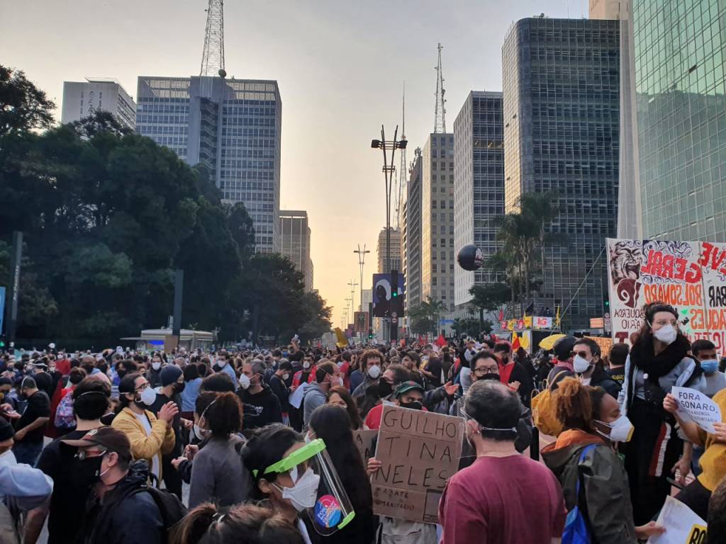 Manifestantes segurando cartazes e portando máscaras ocupam duas vias da Avenida Paulista com pôr do sol ao fundo.
