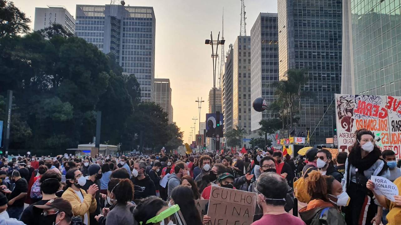 Manifestantes segurando cartazes e portando máscaras ocupam duas vias da Avenida Paulista com pôr do sol ao fundo.
