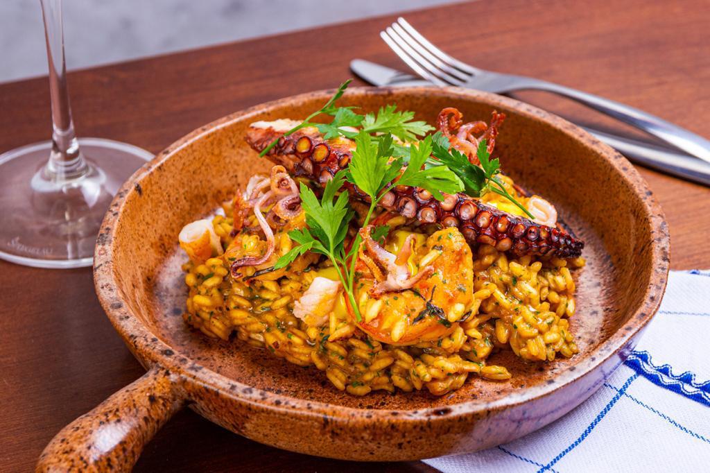 Panelinha alaranjada de pedra com arroz amarelado coberto por camarão, tentáculo de polvo e ramo de salsinha sobre mesa de madeira.