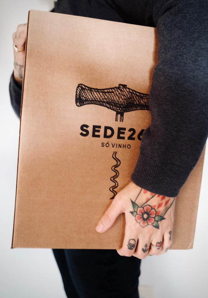 Caixa de papelão com o logo do Sede261 é segurada por pessoa de lado com tatuagens na mão.