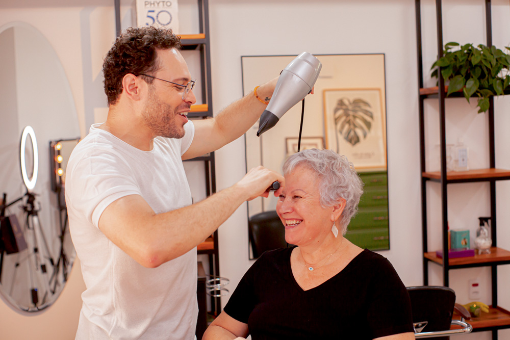 abner-matias-tema-blanco-nosso-louco-amor-diferença-idade-cabeleireiro.jpg abner e telma estão sorrindo enquanto ele cuida do cabelo dela, com um secador em uma mão e uma escova em outra