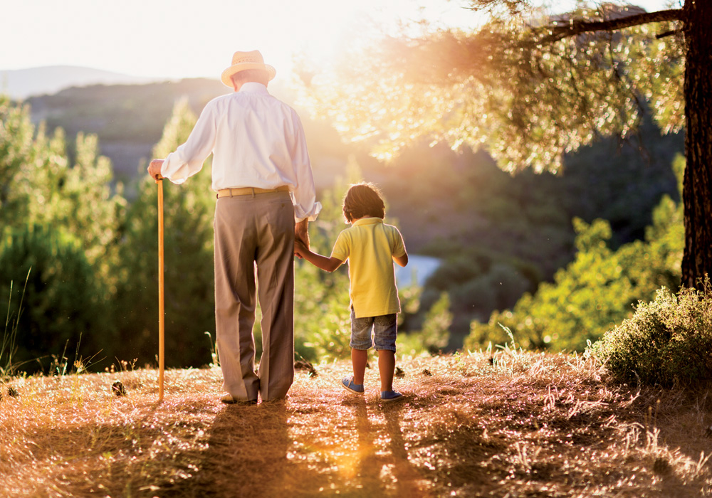 homem apoiado em sua bengala e de mãos dadas com seu neto, andando na grama em um dia ensolarado. eles estão de costas para a foto