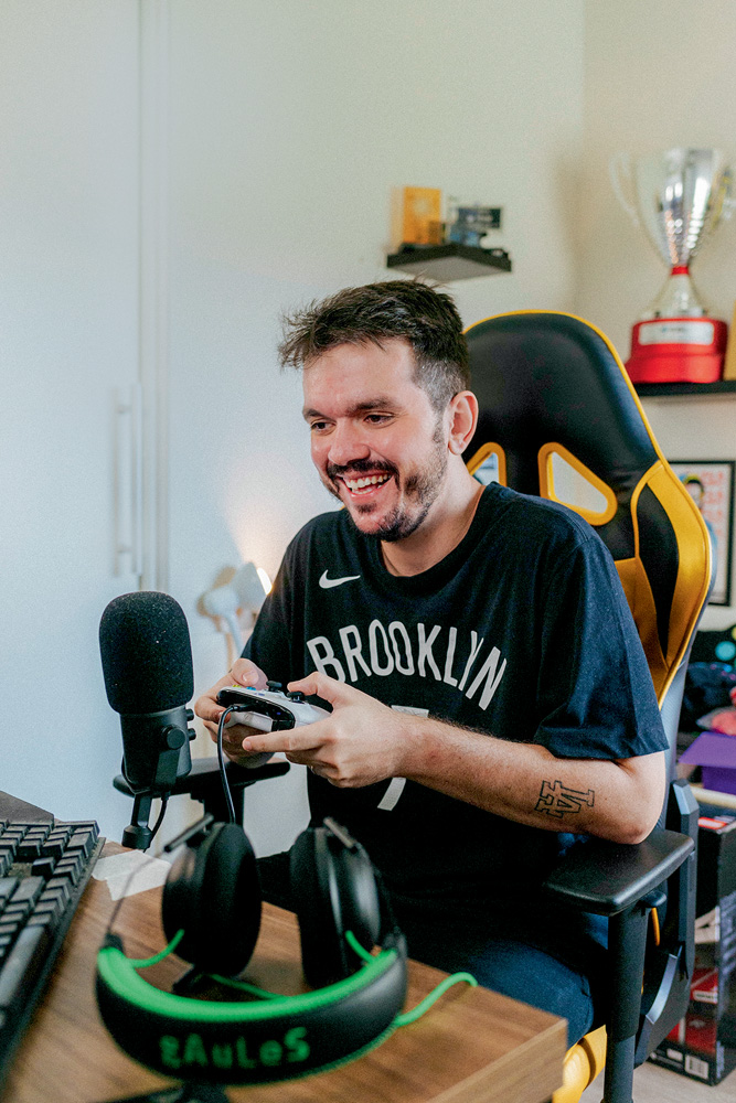 A imagem mostra Gaules, sentado em uma cadeira gamer, rindo para seu monitor e segurando um controle de videogame. Há um microfone profissional na sua frente também.