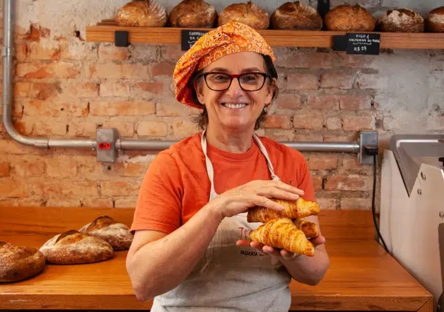 Mulher com avental e pães na mão possa em frente a prateleiras de pães