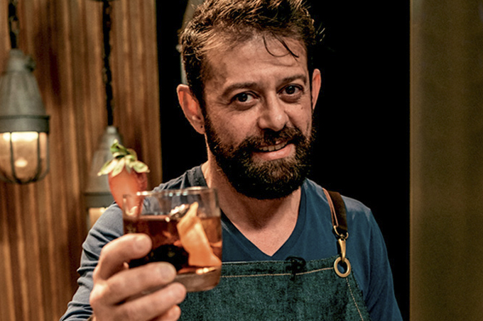 Claudio Galicia oferecendo um drinque de cor âmbar com um morango na borda.