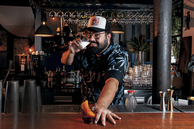 Bartender Diogo Sevilio atrás de balcão de bar segurando um negroni sobre a bancada e tomando um copo de leite.