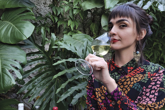 Gabrielli Fleming, sentada à frente de parede de plantas com camisa estampada segurando taça de vinho branco próximo ao nariz.