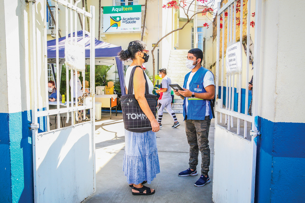 Igor da Silva nos portões de uma unidade básica de saúde segurando tablet e conversando com mulher