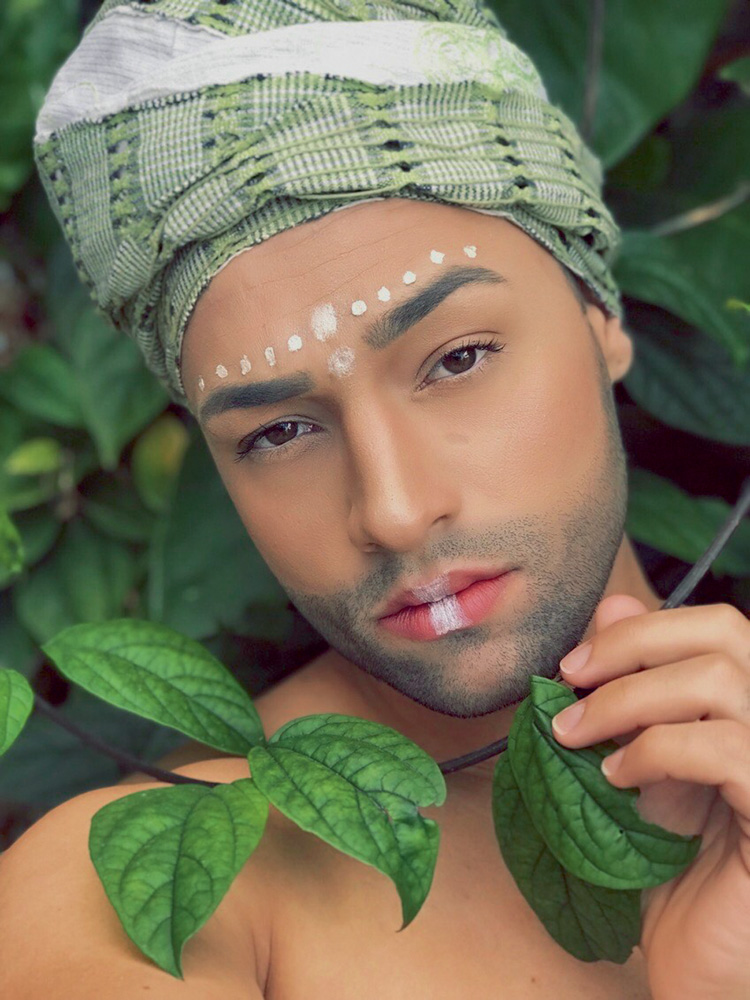 Homem está apoiado em algumas folhas verdes, usando turbante e com algumas pinturas brancas no rosto