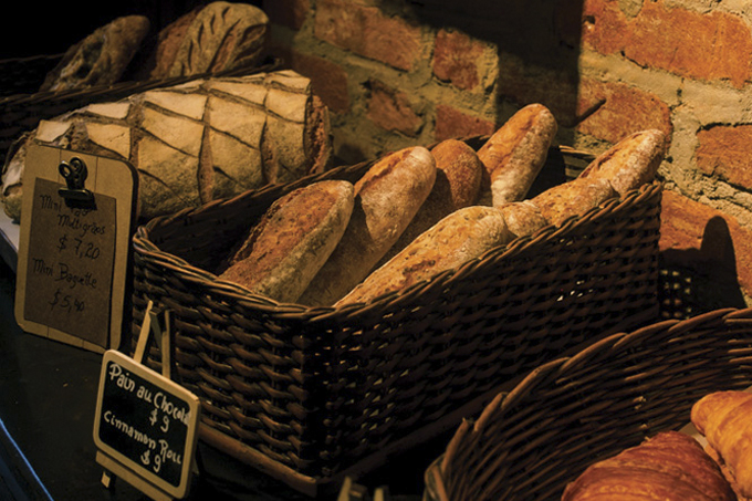 Cestas de pães e croissant sobre bancada de madeira com parede de tijolinhos ao fundo.