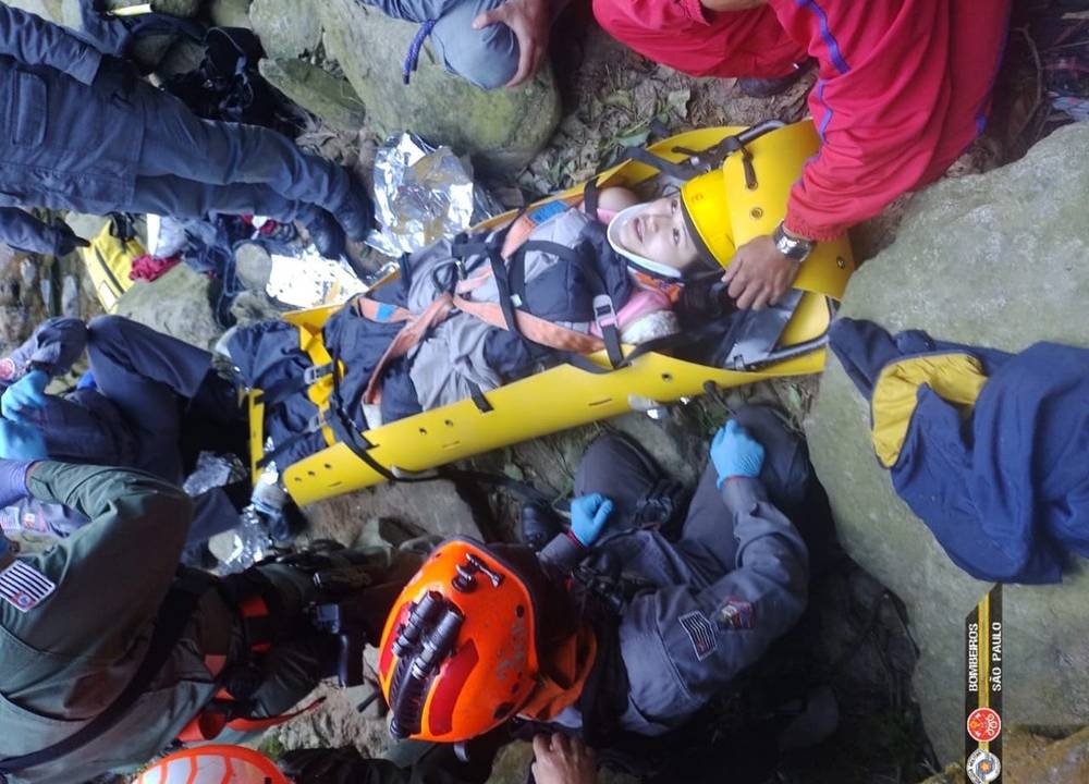 foto tirada de cima para baixo de mulher que sofreu a queda. ela está em uma maca amarela dos bombeiros e está em volta de diversos agentes