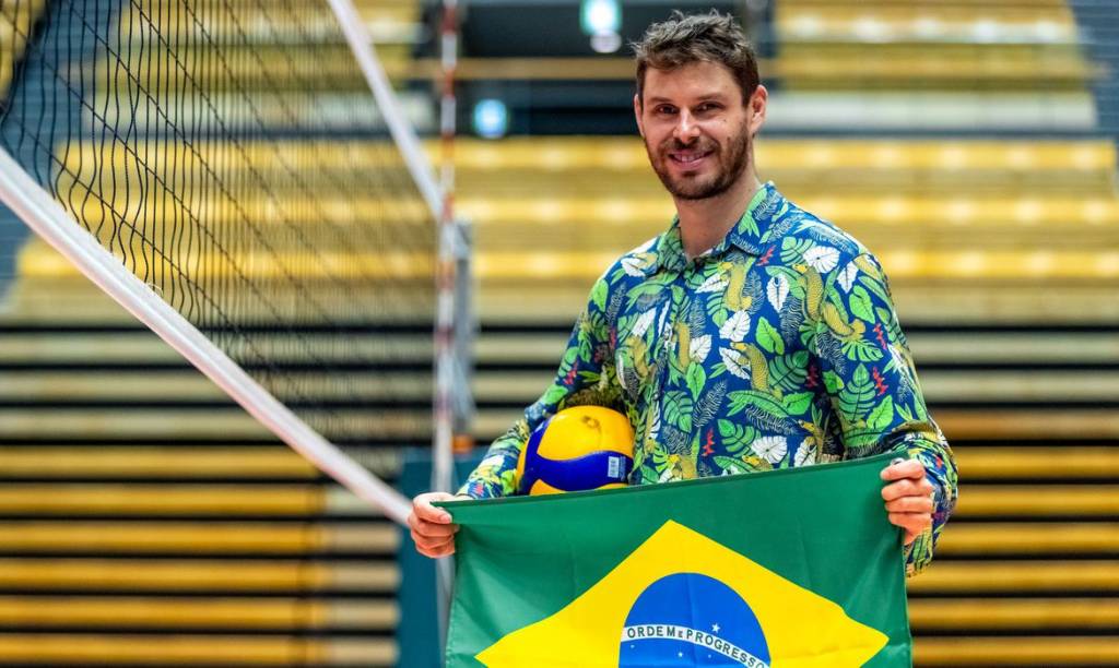 O atleta Bruninho Rezende, do vôlei, segura a bandeira do Brasil, em uma quadra com uma rede de vôlei ao fundo