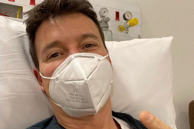 Selfie do apresentador Rodrigo Faro de máscara e camiseta azul deitado em uma cama de hospital.