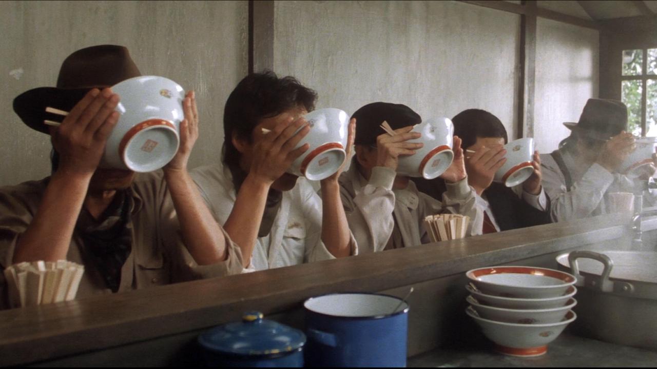 Cena do filme Tampopo: cinco pessoas sentadas atrás de um balcão bebendo algo direto da tigela.