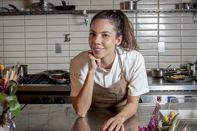 Chef Tássia Magalhães posa apoiada no balcão da cozinha de seu restaurante vestindo avental marrom sobre camisa branca.