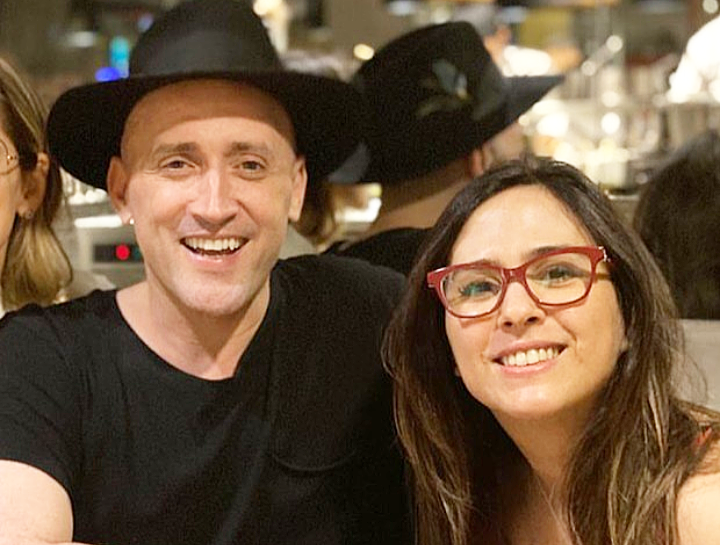 Imagem mostra Paulo Gustavo e Tatá Werneck abraçados, sorrindo. Tatá usa óculos de grau e Paulo, um chapéu