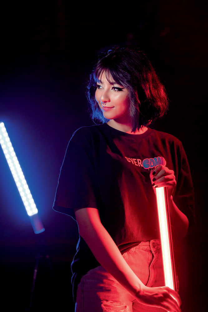 A imagem mostra Taynah, em um estúdio escuro, segurando uma barra de luz vermelha com outra barra azul ao fundo